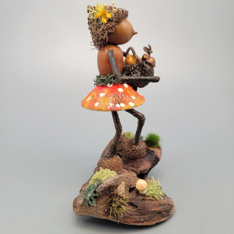 May include: A whimsical figurine of a woodland creature with a mushroom skirt and a basket. The figure is made of natural materials, with a red and white spotted mushroom skirt and a head adorned with a flower. The figurine is standing on a piece of wood.