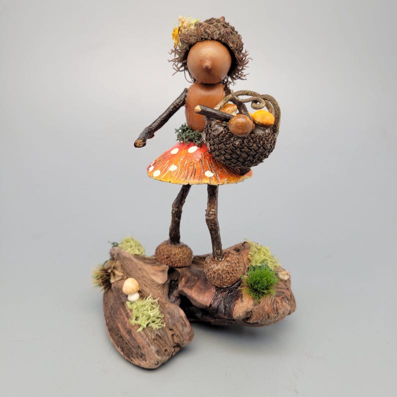 May include: A whimsical figurine of a woodland fairy, crafted from natural materials. The fairy wears a mushroom-shaped skirt with red and white spots and carries a basket of mushrooms. The base is a piece of wood with moss and small mushrooms.