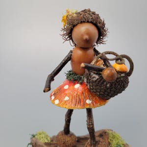 May include: A whimsical fairy figurine crafted from natural materials. The fairy has an acorn body, twig arms and legs, and a mushroom skirt with white polka dots. She carries a basket with miniature mushrooms.