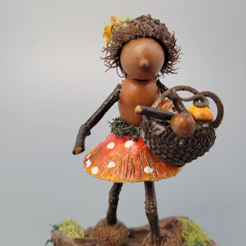 May include: A whimsical fairy figurine crafted from natural materials. The fairy has an acorn body, twig arms and legs, and a mushroom skirt with white polka dots. She carries a basket with miniature mushrooms.