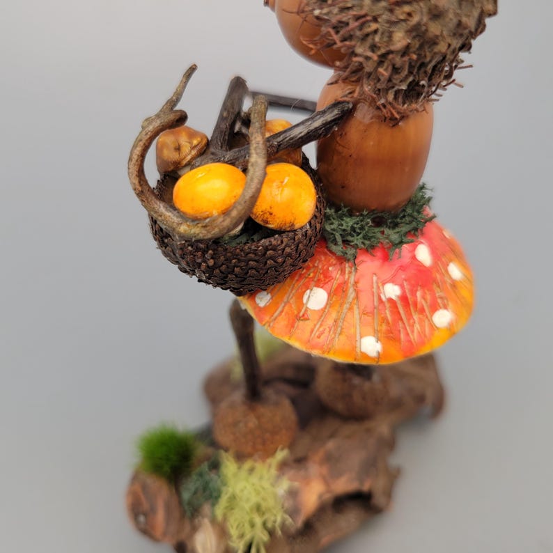 May include: A whimsical figurine of a woodland creature made from natural materials. It carries a basket of orange fruits. The creature wears a red and orange mushroom cap with white spots. The base is made of wood and moss.