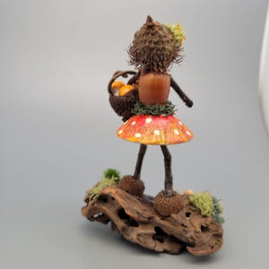 May include: A whimsical handmade figurine of a woodland fairy. The fairy is crafted from natural materials, including an acorn head, a mushroom skirt with red and white spots, and twig legs. It carries a basket of miniature orange mushrooms and stands on a piece of driftwood.