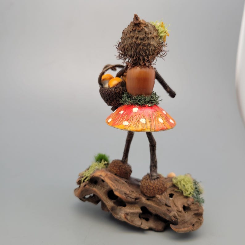May include: A whimsical handmade figurine of a woodland fairy. The fairy is crafted from natural materials, including an acorn head, a mushroom skirt with red and white spots, and twig legs. It carries a basket of miniature orange mushrooms and stands on a piece of driftwood.