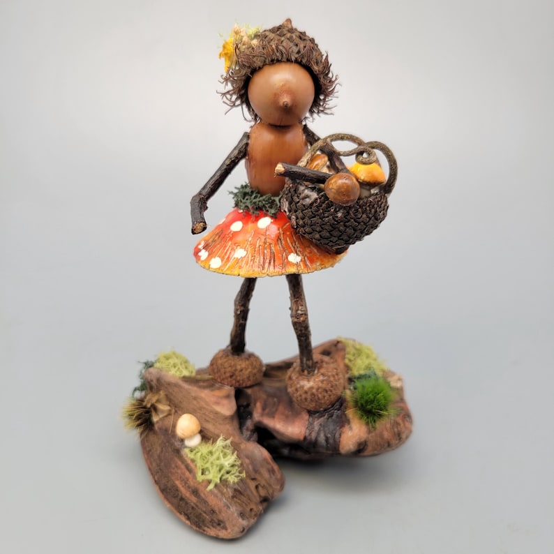 May include: A whimsical handmade figurine of a woodland fairy. The fairy has a brown acorn head, twig arms and legs, and a mushroom skirt. She carries a basket filled with miniature mushrooms and stands on a wooden base with moss.