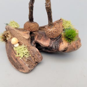May include: A detailed, close-up view of a handcrafted wooden sculpture. The sculpture features a natural wood base with attached elements of moss, a small mushroom, and textured, bulbous forms. The color palette includes browns, greens, and beige.