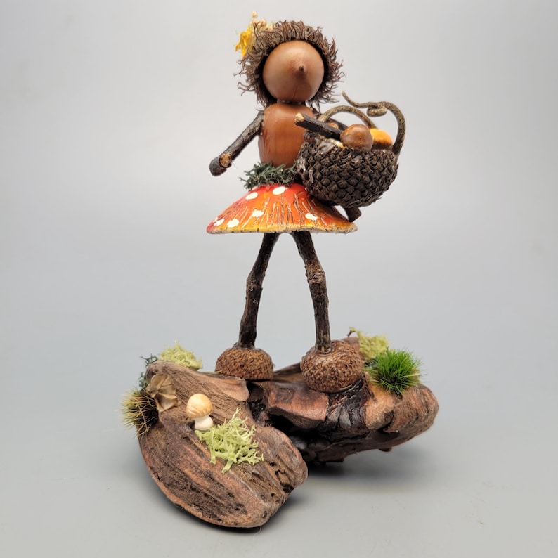 May include: A whimsical wooden figurine of a mushroom fairy. The fairy has a wooden head and arms, a mushroom cap skirt, and twig legs. She carries a basket of mushrooms and stands on a piece of driftwood with moss and small mushrooms.