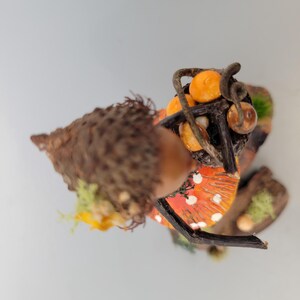May include: A whimsical miniature figurine of a mushroom-like creature carrying a basket of orange fruits. The figurine has a brown acorn cap, a red and white spotted skirt, and is decorated with moss and twigs. The overall theme is fantasy and nature.
