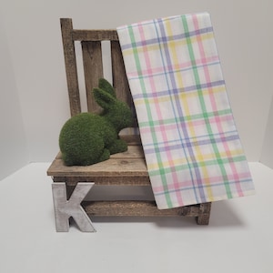 May include: A wooden chair with a green bunny sitting on the seat. A plaid fabric with pink, yellow, green, and blue stripes is draped over the back of the chair. The chair has a wooden letter K on the front.