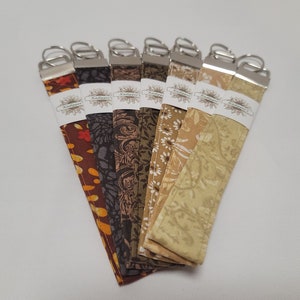 May include: A set of eight fabric keychains with different patterns in shades of brown, green, and beige. Each keychain has a silver key ring and a white label with the text "Kalligraphy" printed on it.
