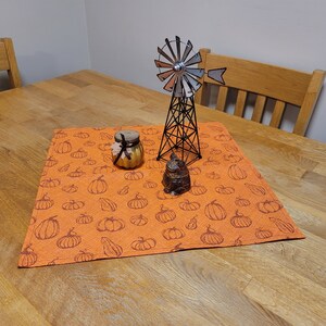 May include: An orange placemat with a repeating pumpkin pattern, a small decorative jar, a metal windmill, and a small decorative container are displayed on a wooden table. The windmill is black with silver blades. The jar has a black ribbon.