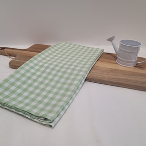 Puede incluir: Una toalla de cocina a cuadros verdes y blancos colocada sobre una tabla de cortar de madera con una pequeña regadera blanca encima.