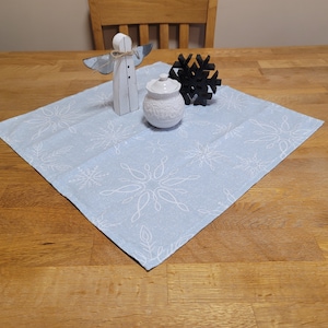 May include: A light blue table runner with a white snowflake pattern. A white ceramic sugar bowl, a wooden angel figurine, and a black wooden snowflake ornament are on top of the runner.