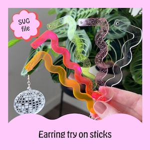 Puede incluir: Una colección de palos ondulados y coloridos para probar pendientes, en tonos rosa, naranja, verde y transparente. Un pendiente de bola de discoteca cuelga de uno de los palos. La imagen incluye el texto "SUG file" y "Earring try on sticks".