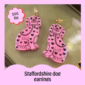 Op de afbeelding: Roze Staffordshire-dog oorbellen met zwarte hartaccenten en een kettinghalsbandontwerp. De oorbellen hebben goudkleurige pinnen. De tekst "SUG file" staat in een roze wolk en de tekst "Staffordshire dog earrings" staat onderaan, tegen een lichtgroene achtergrond.