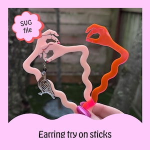 May include: Two plastic earring try-on sticks. One stick is pink and has a hand holding a silver dove earring. The other stick is orange and has a hand with a wavy design. The text "SVG file" is in a white cloud.