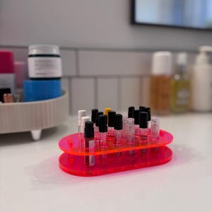 May include: A pink acrylic organizer with two tiers holds a collection of small glass bottles with black caps. The bottles are arranged in rows and are filled with a clear liquid.