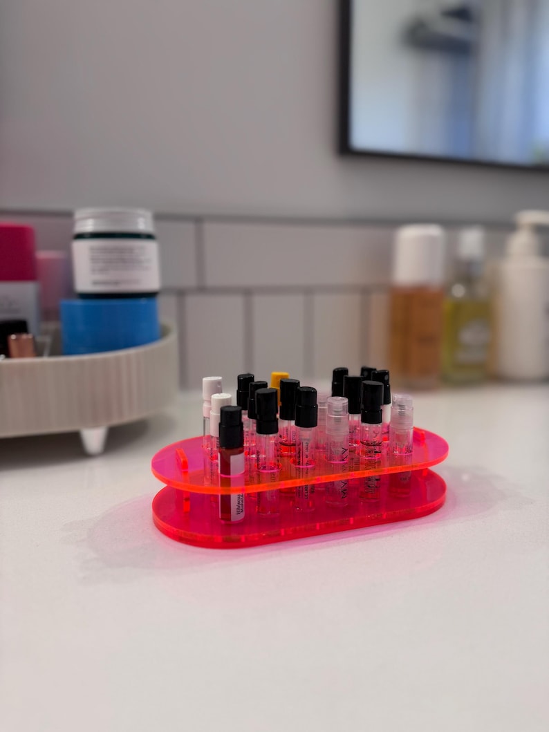 May include: A pink acrylic organizer with two tiers holds a collection of small glass bottles with black caps. The bottles are arranged in rows and are filled with a clear liquid.