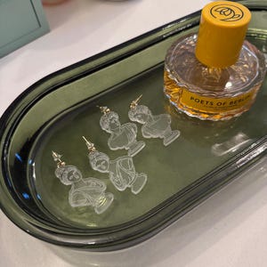 May include: A green oval-shaped tray with four pairs of clear acrylic earrings featuring a bust of a woman. The earrings are on a green tray with a bottle of perfume labeled "POETS OF BERLIN" in the background.