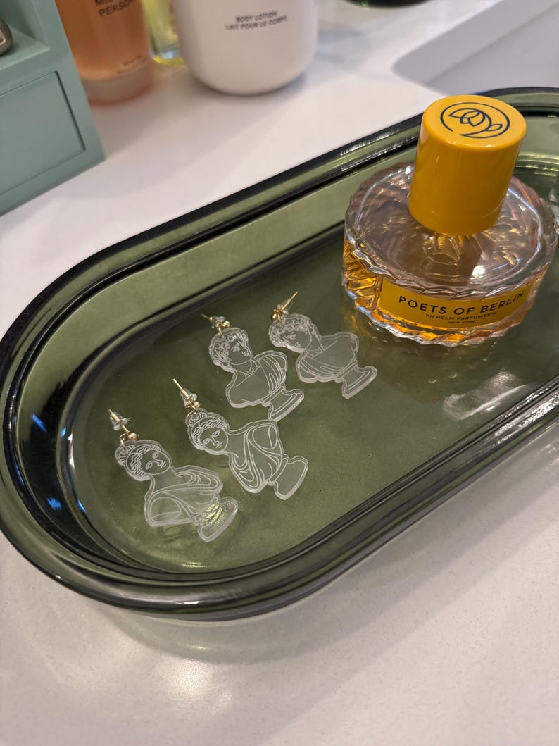 May include: A green oval-shaped tray with four pairs of clear acrylic earrings featuring a bust of a woman. The earrings are on a green tray with a bottle of perfume labeled "POETS OF BERLIN" in the background.