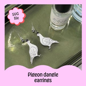 May include: Silver dove dangle earrings with a white outline. The earrings are on a green surface with a bottle of clear liquid in the background.