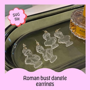 May include: Four pairs of clear acrylic dangle earrings featuring a Roman bust design. The earrings are displayed on a gray tray with a bottle of perfume in the background. The text "SVG file" is visible in a green cloud shape.