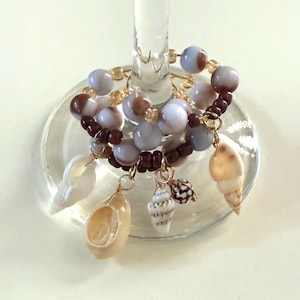 May include: A wine glass charm featuring a beaded ring with light blue, brown, and gold beads. The charm is adorned with several seashells of varying shapes and sizes, suspended by gold wire.