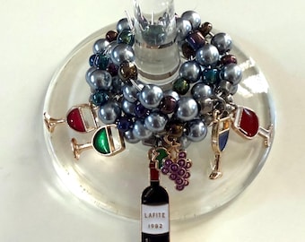May include: A wine glass charm featuring a cluster of gray beads and wine-themed charms. Charms include miniature wine glasses with red, green, and blue accents, a wine bottle with "LAFITE 1992" text, and a bunch of purple grapes.