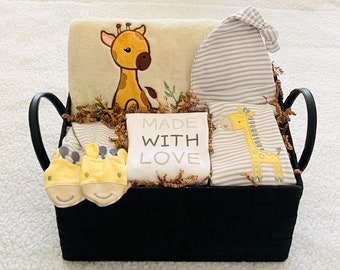 May include: A black basket filled with baby items, including a blanket, a hat, a onesie, and a pair of booties. The items are decorated with a giraffe pattern and the words "Made with Love".