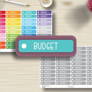 Puede incluir: Una hoja de pegatinas de presupuesto de colores con la palabra "PRESUPUESTO" impresa en cada pegatina. Las pegatinas están dispuestas en filas y columnas sobre una superficie de madera.