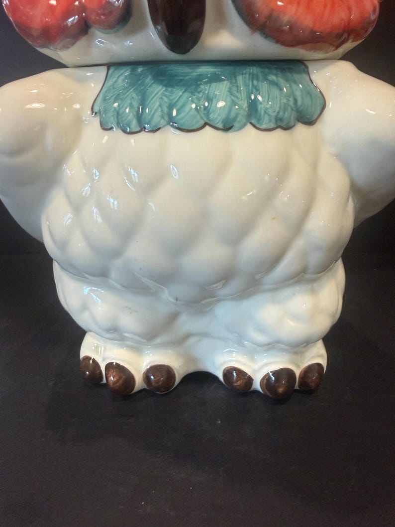 May include: A ceramic owl-shaped cookie jar. The jar is primarily white with textured detailing, a turquoise collar, and brown feet. The owl's head features red and brown accents.
