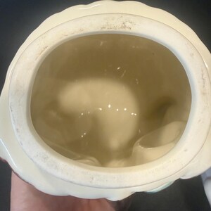 May include: A vintage ceramic cookie jar with a cream-colored exterior and a scalloped rim. The interior is a light beige color. The jar has a round opening and appears to be in good condition.