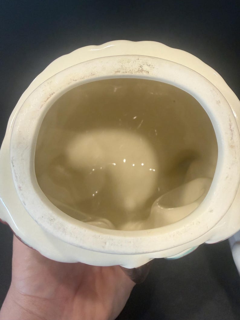 May include: A vintage ceramic cookie jar with a cream-colored exterior and a scalloped rim. The interior is a light beige color. The jar has a round opening and appears to be in good condition.