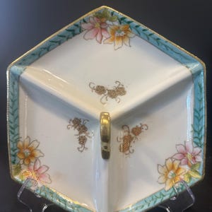 May include: A hexagonal, divided serving dish with a gold handle. The white porcelain dish features a teal border with gold trim and floral designs in pink, yellow, and orange. The dish is supported by a clear stand.
