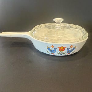 May include: A white, handled casserole dish with a clear glass lid. The dish features a colorful floral and bird design in blue, orange, and yellow. The handle is integrated into the dish's design.