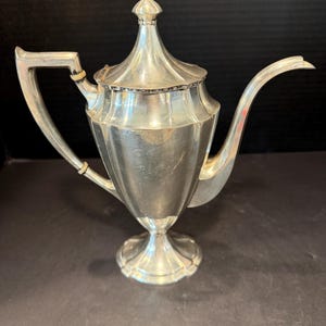 May include: A silver-toned metal coffee pot featuring a tall, tapered body with vertical fluted panels, a pointed lid with a finial, a curved handle with two light-colored rings, and a long, slender spout extending from the side on a pedestal base.