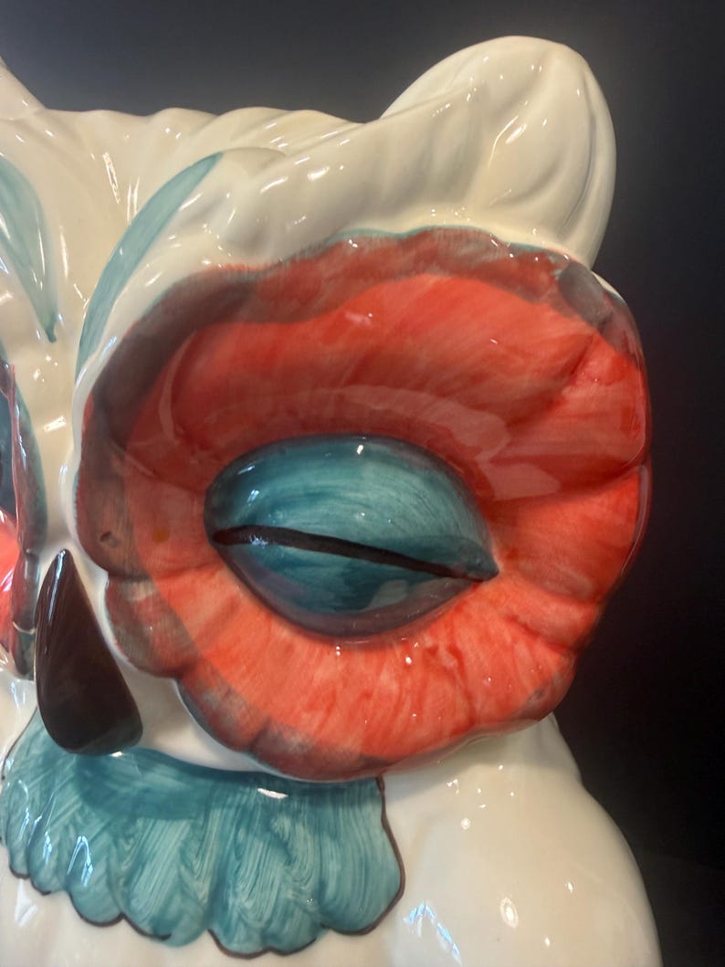 May include: A ceramic owl figurine with a glossy finish. The owl features a white body, red and teal accents, and a large, detailed eye. The eye is a vibrant teal color with a black slit pupil. The owl's design is whimsical and decorative.