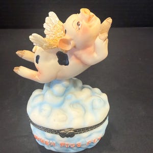 May include: A whimsical figurine of a pig with angel wings, mid-flight above a cloud-shaped trinket box. The box is white and blue with the phrase "WHEN PIGS FLY" in orange lettering. The pig has black spots.