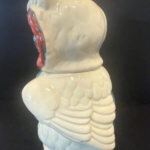 May include: A vintage ceramic owl cookie jar. The jar is primarily white with red and teal accents. The owl has large, detailed wings and a rounded body. The lid is shaped like the owl's head, with a pointed top and a detailed face.