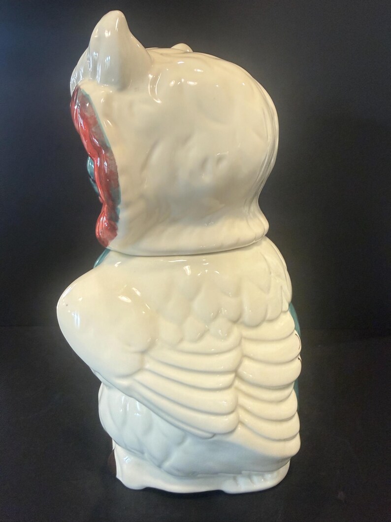 May include: A vintage ceramic owl cookie jar. The jar is primarily white with red and teal accents. The owl has large, detailed wings and a rounded body. The lid is shaped like the owl's head, with a pointed top and a detailed face.