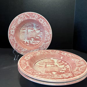 May include: Set of three pink and white decorative plates. Each plate features a central landscape scene with a bridge and trees, surrounded by a floral border. The plates are stacked on a dark surface.