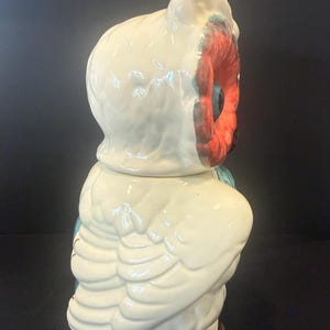 May include: A ceramic owl-shaped cookie jar. The jar is primarily off-white with red, blue, and brown accents. The owl has large, detailed wings and a textured surface. The jar is designed to resemble a stylized owl.