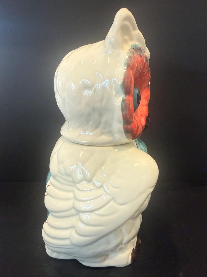 May include: A ceramic owl-shaped cookie jar. The jar is primarily off-white with red, blue, and brown accents. The owl has large, detailed wings and a textured surface. The jar is designed to resemble a stylized owl.