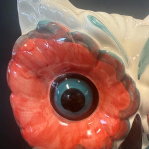 May include: Close-up of a ceramic owl figurine. The owl features a large, red and white eye with a blue iris and a black pupil. The owl's head is white with red and blue accents. The figurine has a glossy finish.