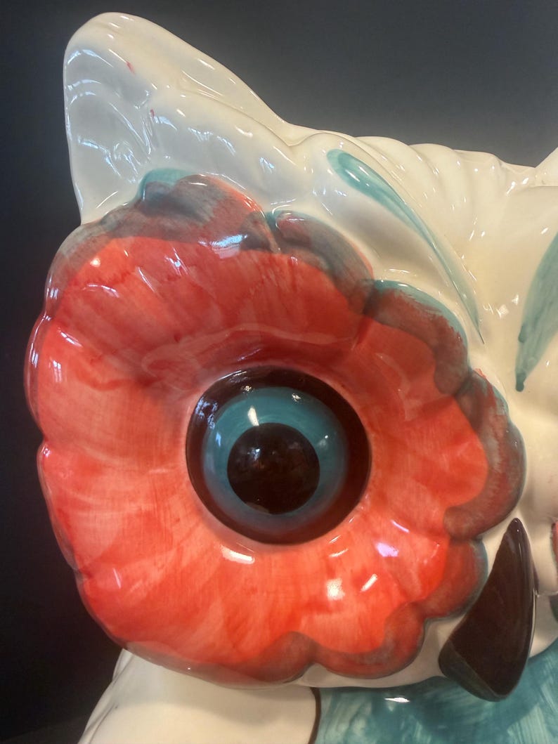 May include: Close-up of a ceramic owl figurine. The owl features a large, red and white eye with a blue iris and a black pupil. The owl's head is white with red and blue accents. The figurine has a glossy finish.