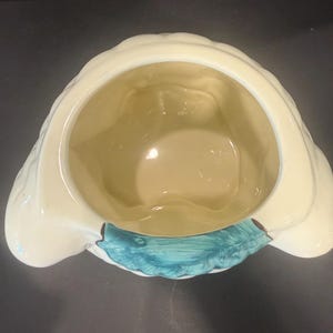 May include: A cream-colored ceramic container with a scalloped rim and a teal accent. The interior is a light yellow. The container has a unique shape, possibly for decorative or functional use.