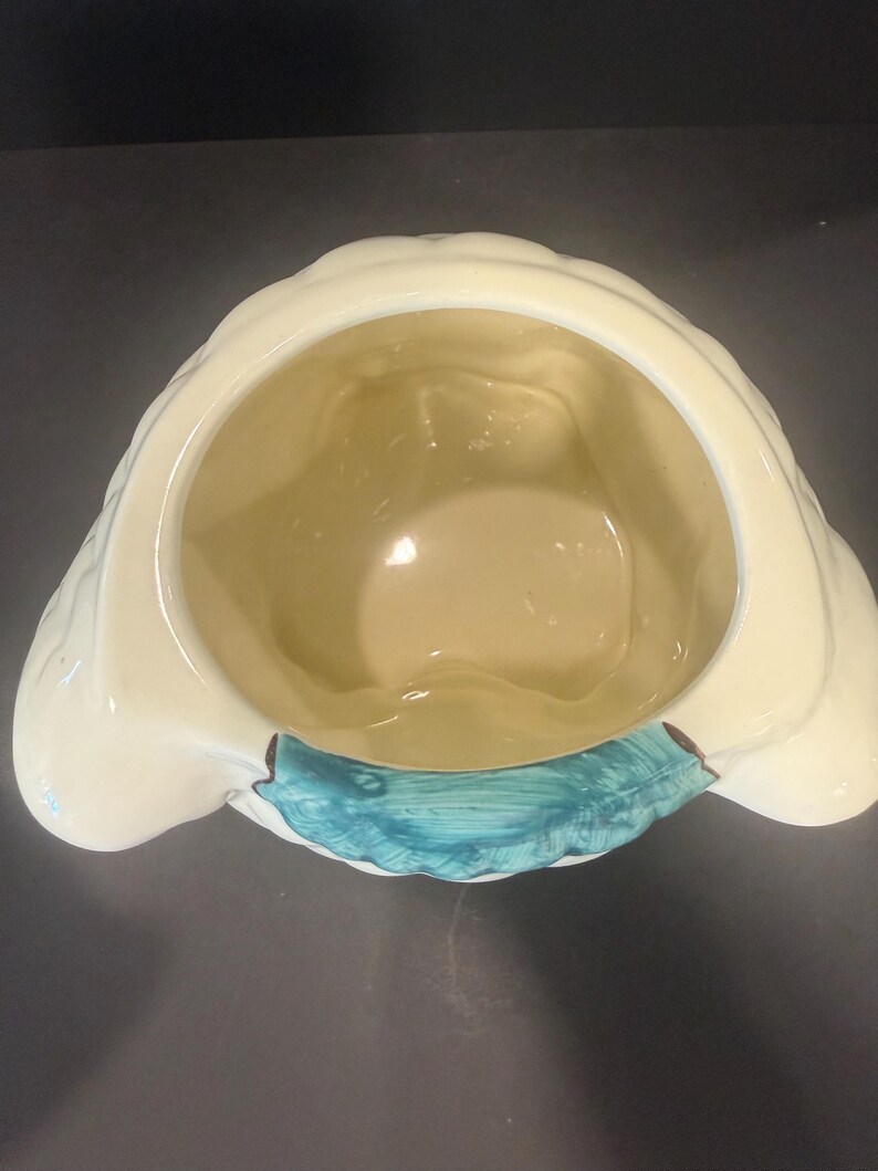 May include: A cream-colored ceramic container with a scalloped rim and a teal accent. The interior is a light yellow. The container has a unique shape, possibly for decorative or functional use.