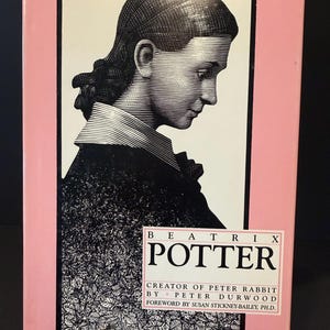 May include: A book cover featuring a black and white illustration of a woman in profile, set against a pink background. The title "BEATRIX POTTER" is prominently displayed, with author and foreword details below. The book is displayed on a clear stand.
