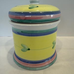 May include: A ceramic canister with a lid. The canister is yellow with blue, pink, and green stripes. The lid has a small knob and is also yellow with stripes. Green leaf designs are painted on the canister.