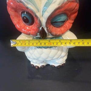 May include: A ceramic owl figurine with a white body, red and blue accents, and a brown beak. The owl's eyes are blue and red. The figurine is approximately 6 inches tall.