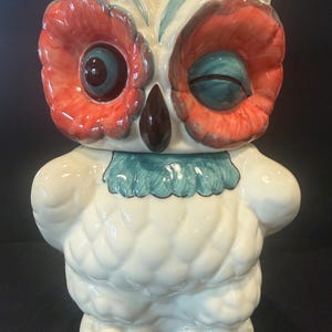May include: A ceramic owl-shaped cookie jar. The owl has a white body, red and teal eyes, and a teal collar. The beak is brown. The jar is a decorative kitchen item.
